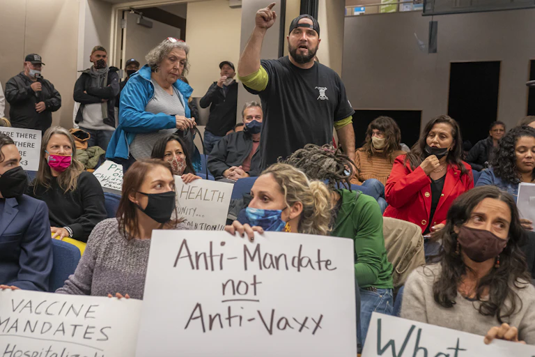 A angry man stands up and points in a crowd filled with people, most of whom are wearing masks and holding protest signs.