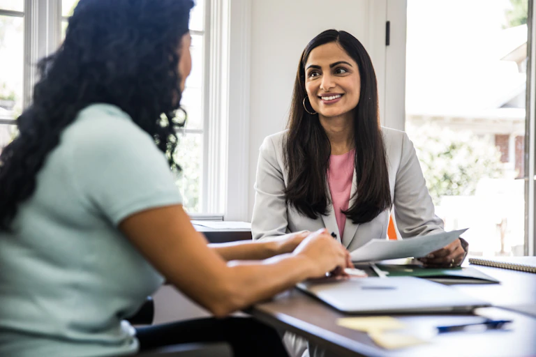 Two women talk at a conference table while holding documents.