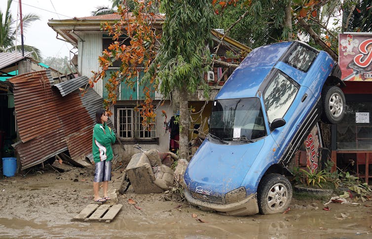 Uma mulher está de braços cruzados, olhando para um carro apoiado no nariz contra uma empresa após o tufão.
