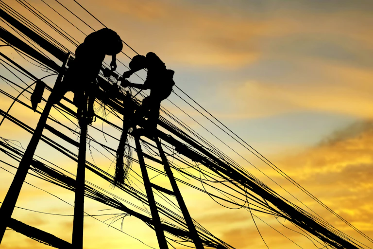 Low angle view of silhouette men working on power cables against orange sky.