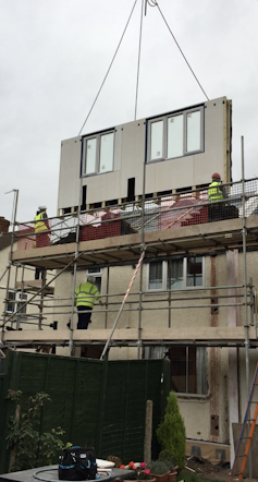 A crane lowers down a wall and windows over an existing house.