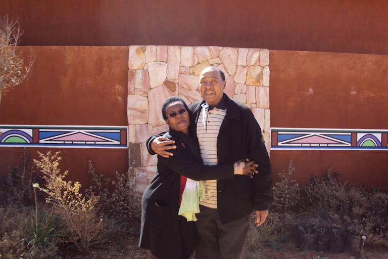 A man and woman pose together for a photograph against a stone wall with traditional African murals painted on either side of it. She holds him and he has an arm around her.
