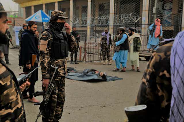 Taliban fighters in combat fatiguesa dnd COVID masks with the corpse of a suicide bomber shot attempting to detonate a bomb. In the background are Afghan citizens.