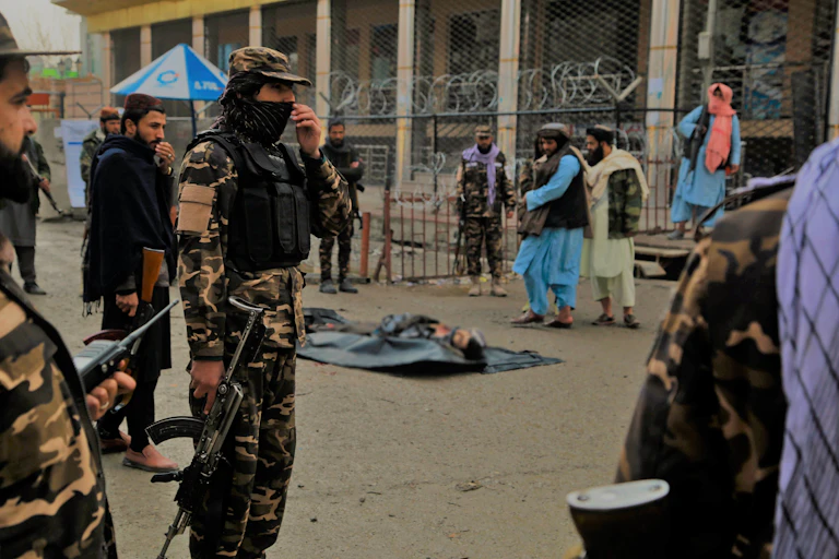 Taliban fighters in combat fatiguesa dnd COVID masks with the corpse of a suicide bomber shot attempting to detonate a bomb. In the background are Afghan citizens.