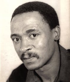 A black and white image of a young man, an earnest expression on his face, hair short and sporting a moustache.
