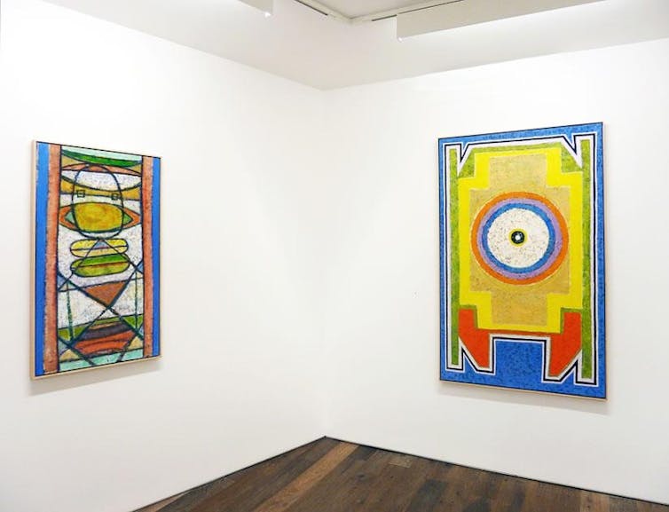 Two brightly colored paintings hanging on the wall of an art gallery.  They feature circular and angular shapes in yellows, reds and blues.