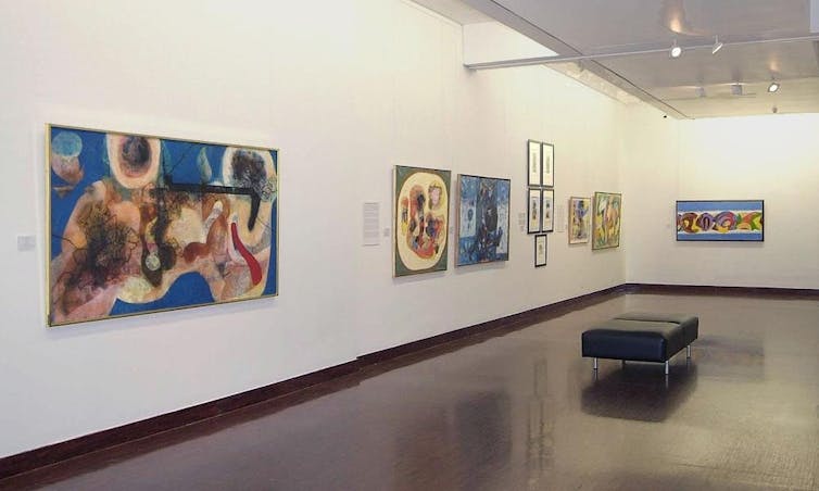 Numerous paintings hanging on two art gallery walls, featuring fluid, abstract forms and shapes in bright colours.