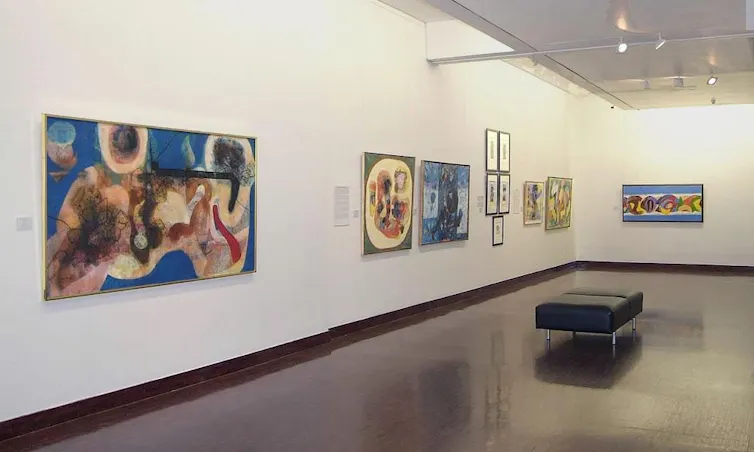 Numerous paintings hanging on two art gallery walls, featuring fluid, abstract forms and shapes in bright colours.