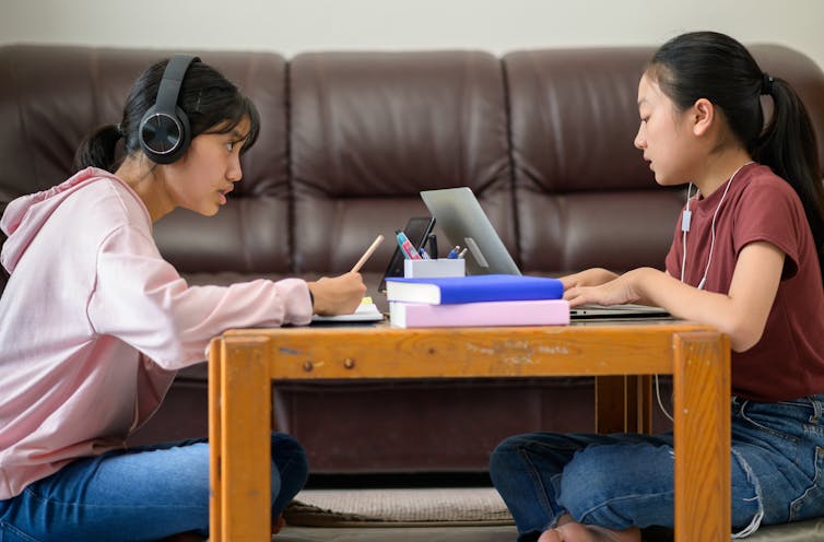 Two teenage girls study at home with laptops and headphones.