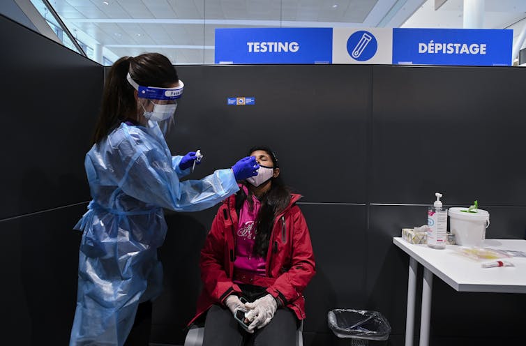 A woman in a red jacket gets a PCR test from a nurse in wearing protective personal equipment. A sign reading testing is over their heads.