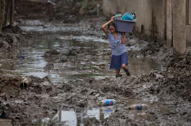 A woman wades through mud and water, carrying a plastic tub full of household items on her shoulder. 