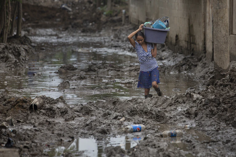 A woman wades through mud and water, carrying a plastic tub full of household items on her shoulder.