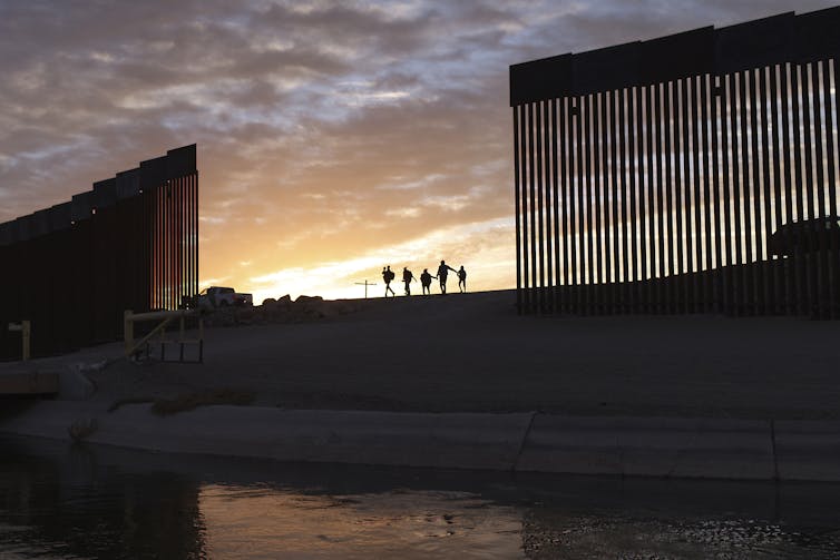 A backlit photo of the border wall with the silhouettes of six people walking along a ridge with the river below.