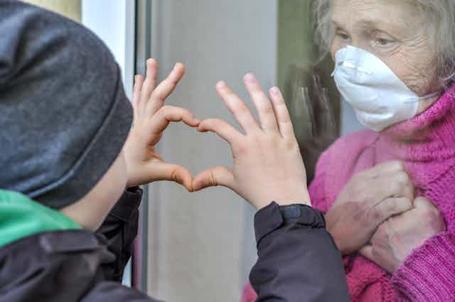 A woman stands in a window wearing a pink sweater with her hands crossed over her chest, she's wearing a mask. A boy presses his hands up against the window, making a heart sign.