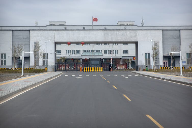A road leads up to a building where police officers stand, the building looks like a large prison, the walls are cement and tall.