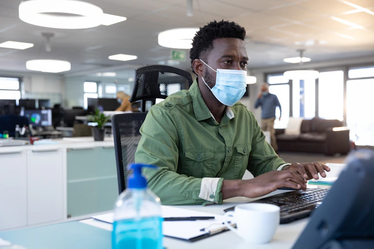 A man wearing a green shirt sits at a computer wearing a mask. He is in an office, he looks concerned.
