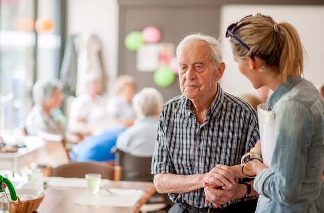 Elderly man in plaid shirt holding the hand of a female caregiver in a nursing home setting.