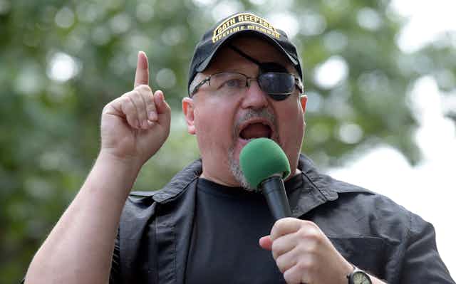 Oath Keepers founder Stewart Rhodes wearing a eyepatch and baseball hat speaks into a microphone while holding his finger aloft.