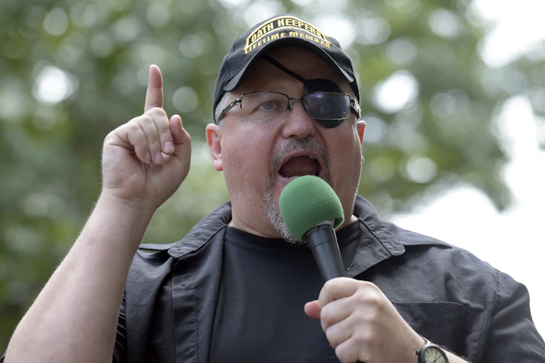 Oath Keepers founder Stewart Rhodes wearing a eyepatch and baseball hat speaks into a microphone while holding his finger aloft.