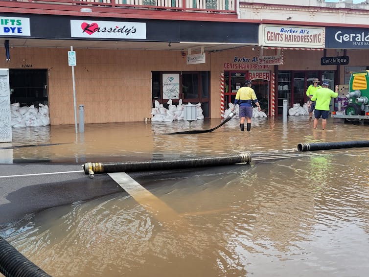 Men standing in flooded street in front of stores, with pumping equipment.