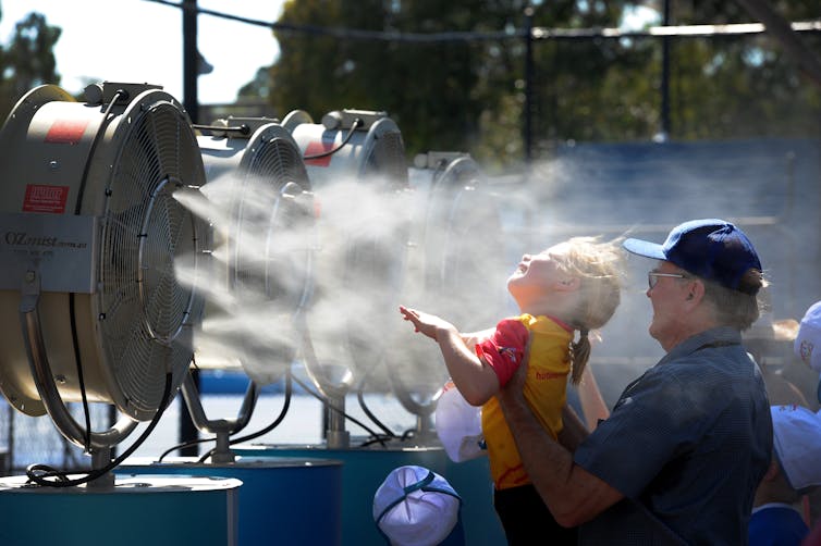 Man holds blond child in front of big fan with cooling mist being blown out.