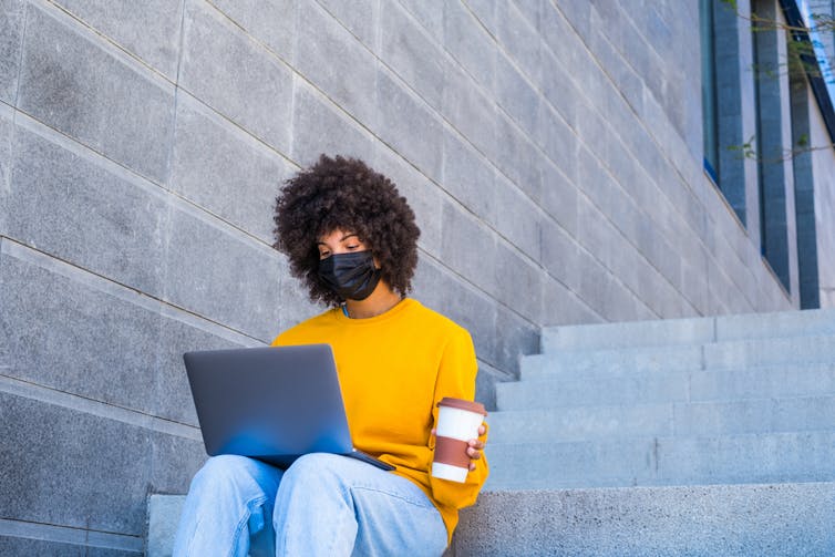 Woman wearing mask, sitting on steps, looking at laptop, holding takeaway coffee cup