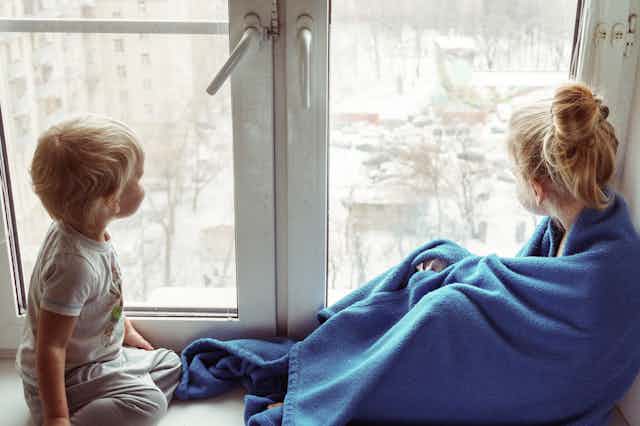 Two children look out a window, one in a blanket.