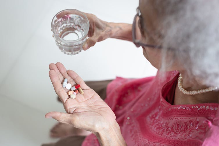 Elderly woman holding multiple pills in her left hand, and a glass of water in her right hand.