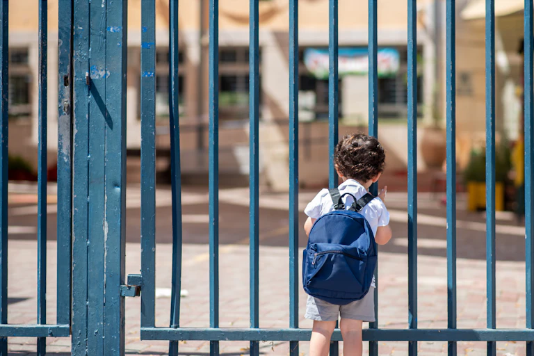 Un niño con mochila asomado a la puerta cerrada de un colegio.
