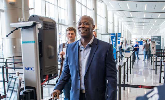 A dark-skinned man in business attire smiles as he stands in front of an electronic device in an airport terminal