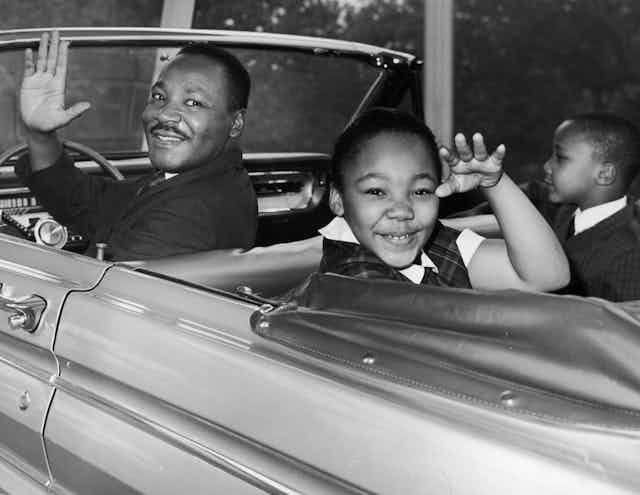 Martin Luther King Jr. waves from the driver's seat of a 1960s-era car with his children in the back seat.