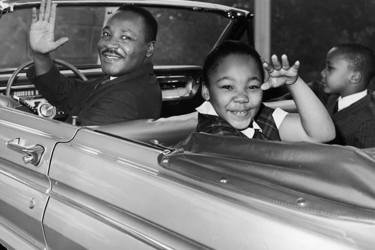 Martin Luther King Jr. waves from the driver's seat of a 1960s-era car with his children in the back seat.