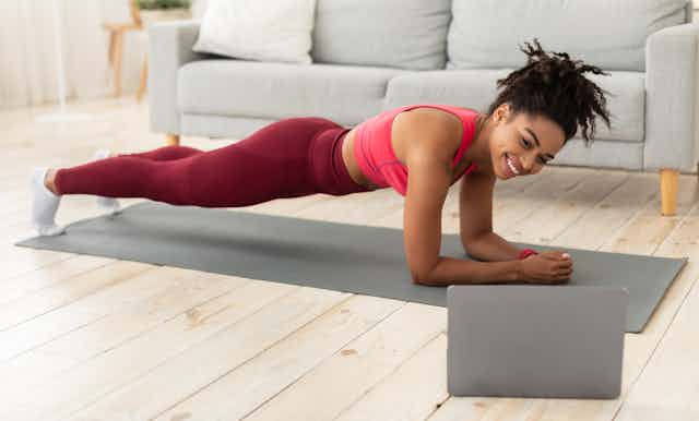 A woman in athletic wear doing a plank on a yoga mat while looking at her laptop screen