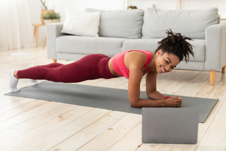 A woman in athletic wear doing a plank on a yoga mat while looking at her laptop screen