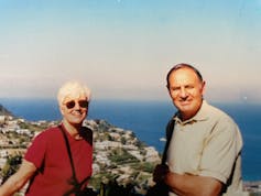 Frank Piccolo and his wife, Theresa, standing near each othe, on vacation, with a hillside village and the sea behind them.