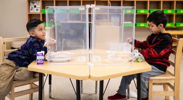 Two boys are seen seated at a table with dividers eating food at school.