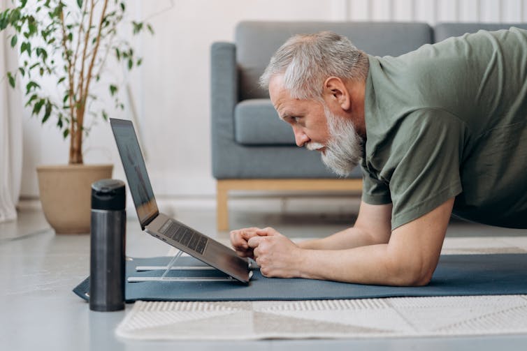 An older man with a beard on a yoga mat looking at a laptop screen