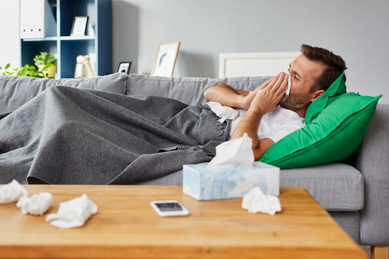 A sick man lies on a sofa at home, blowing his nose and the coffee table is littered with used tissues