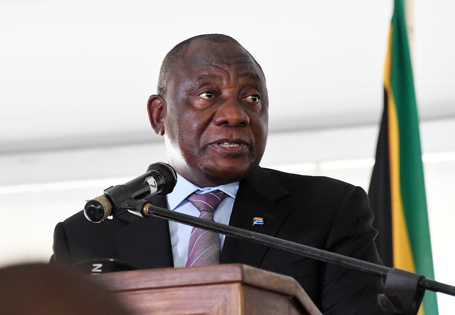 A man wearing a shirt, tie and jacket talks into a microphone at a podium, with the fla of South Africa in the background.