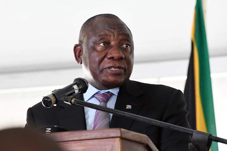 A man wearing a shirt, tie and jacket talks into a microphone at a podium, with the fla of South Africa in the background.