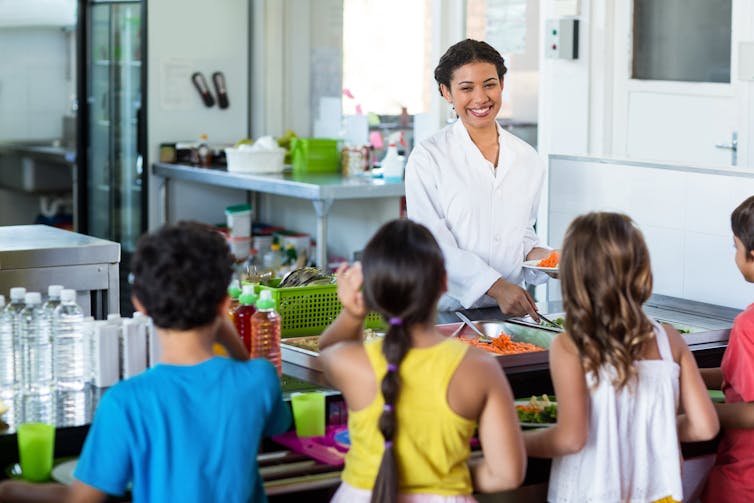 Children stand with trays in a cafeteria and a person serves food.