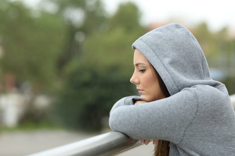 Sad young woman wearing a grey hooded jumper on a balcony stares into the distance.