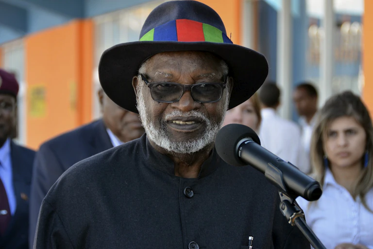 A man with a grey beard and a hat with a multicoloured band on it, in a black shirt and sunglasses, speaks into a microphone.