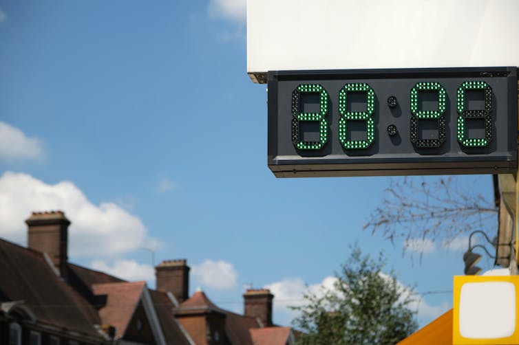A digital thermometer displays 38°C in front of a row of terraced houses.