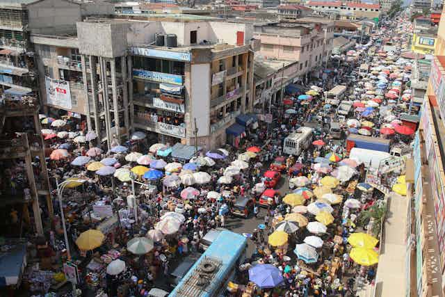 An aerial view of an urban scene, umbrellas crowded alongside a rode indicating market stalls, people thronging everywhere.