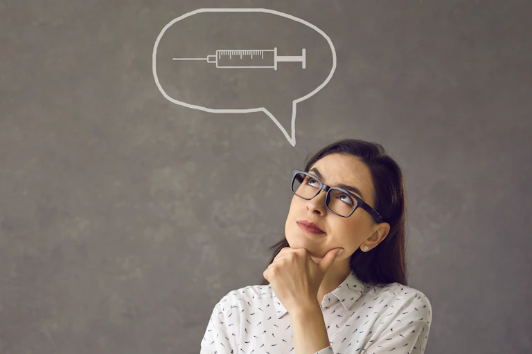 A woman wearing glasses in a thinking pose with a syringe in a thought bubble over her head