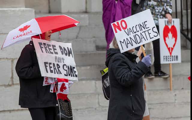 Two people hold signs at a protest; one reads no vaccine mandates. the other carries a red and white umbrella with a maple leaf on it carrying a sign that says freedom not fear some.