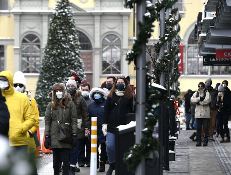 People wearing masks stand in an outdoor lineup with a Christmas tree in the background.