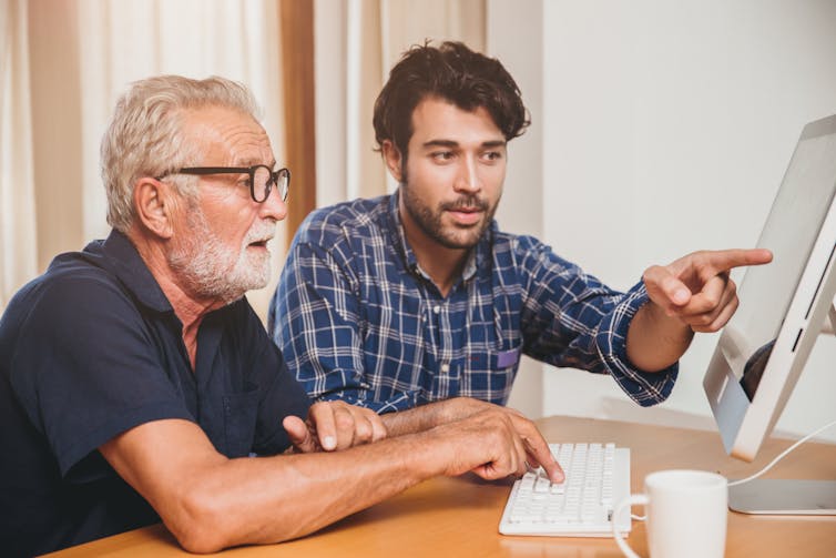 an older man sits in front of a laptop while a younger man is pointing at the screen