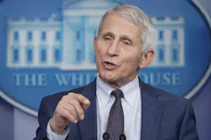 A grey-haired man in a suit gestures as he talks with the White House insignia on the wall behind him.